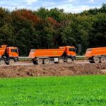 Three orange dump trucks parked at an outdoor construction site beside green grass fields.