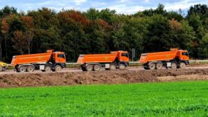 Three orange dump trucks parked at an outdoor construction site beside green grass fields.